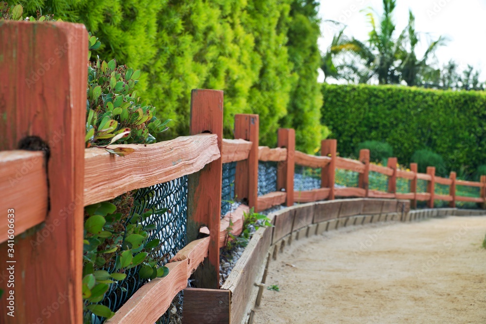 Wooden fence along pathway. Outdoor walk in a countryside. Horizontal rural landscape. Green bushes and sandy soil. Nobody outside.
