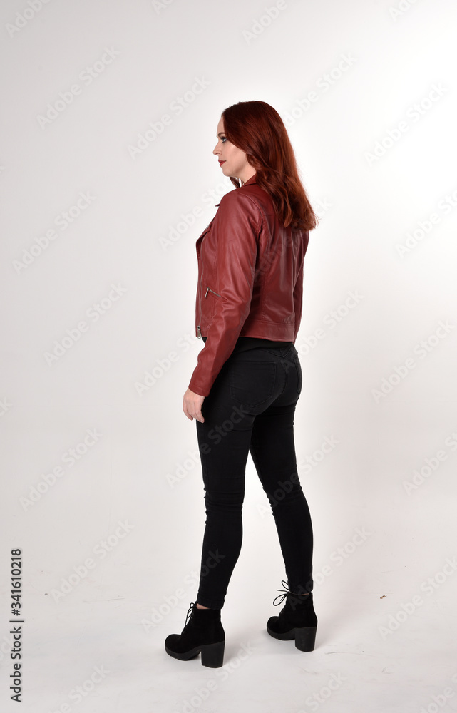 Portrait of a pretty girl with red hair wearing black jeans and boots with leather jacket. full length standing pose with back to the camera a studio background. Portrait of a pretty girl with red hair wearing black jeans and boots with leather jacket. full length standing pose with back to the camera a studio background.