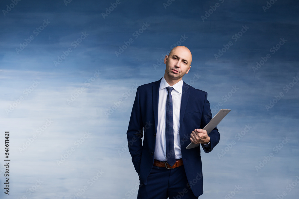 Young bald male businessman in a blue suit holds a folder on a blue background. Young bald male businessman in a blue suit holds a folder on a blue background.
