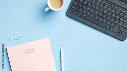 Modern office desk table with laptop keyboard, cup of coffee, paper notebook, pen, glasses on blue background. Writer or blogger workspace, flat lay, top view. Business and technology concept