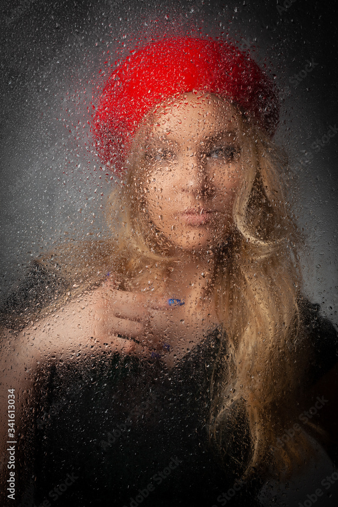 Blonde girl in a red beret behind wet glass on a gray background.