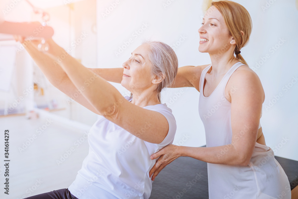Blond female instructor helping, consulting old age grey hair beautiful woman workout pilates on reformer practice in pilates studio, working out indoor, correcting beginners, frehabilitation concept. Blond female instructor helping, consulting old age grey hair beautiful woman workout pilates on reformer practice in pilates studio, working out indoor, correcting beginners, frehabilitation concept.