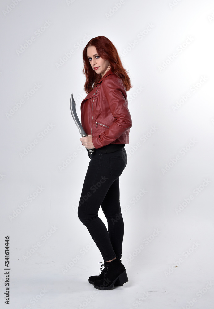 Portrait of a pretty girl with red hair wearing black jeans and boots with leather jacket. full length standing pose holding knife a studio background. Portrait of a pretty girl with red hair wearing black jeans and boots with leather jacket. full length standing pose holding knife a studio background.