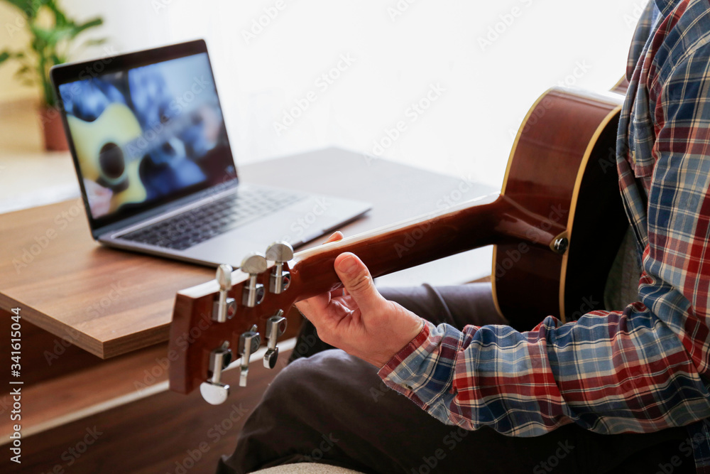 Fototapeta premium Music college hipster student in checkered shirt practicing acoustic guitar exercise, reading notes from laptop. Man taking an online musical courses at home during quarantine. Background, close up