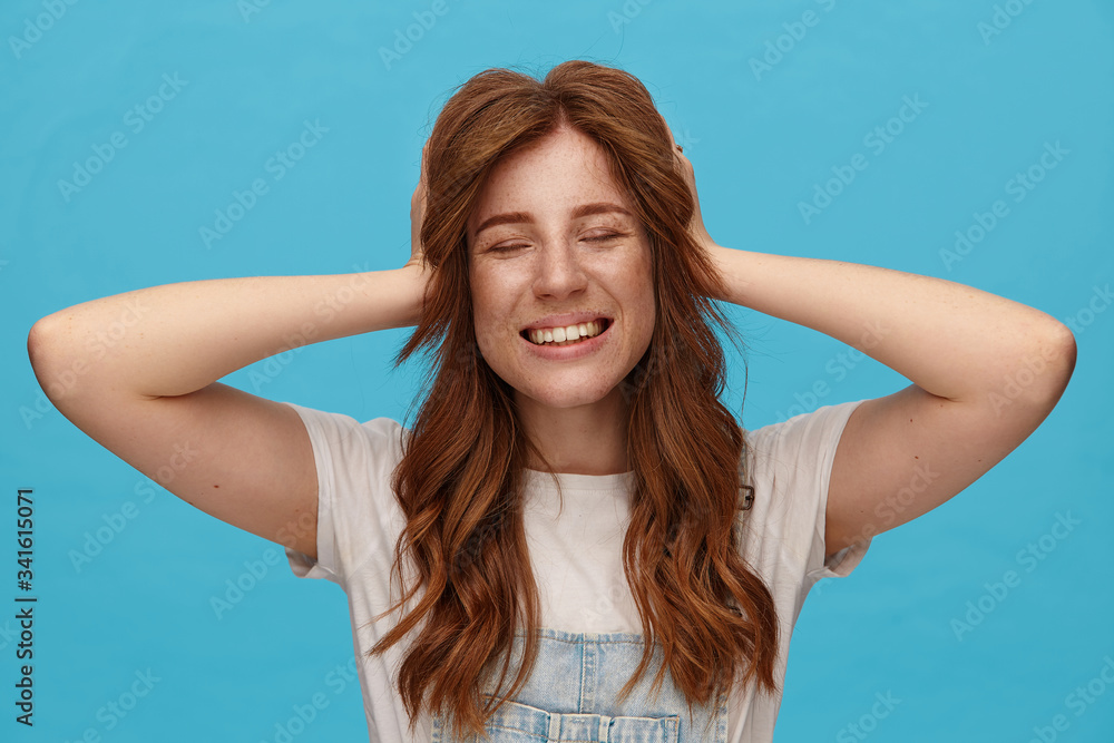 Pleasant looking young lovely redhead female holding her head with raised hands and smiling cheerfully with closed eyes, standing over blue background in casual wear Pleasant looking young lovely redhead female holding her head with raised hands and smiling cheerfully with closed eyes, standing over blue background in casual wear