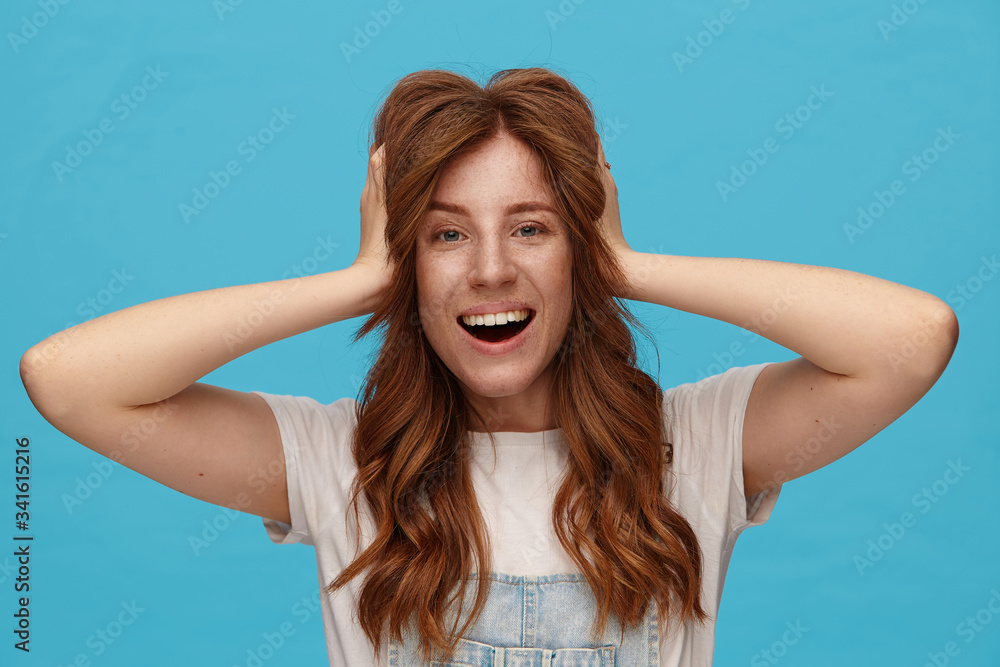 Joyful young attractive woman with foxy wavy hair holding raised hands on her head and looking happily at camera with wide smile, isolated over blue background