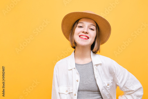Pretty smiling tourist girl isolated on yellow background, looking at camera and smiling. Lady in hat and summer clothes stands on orange background. Copy space