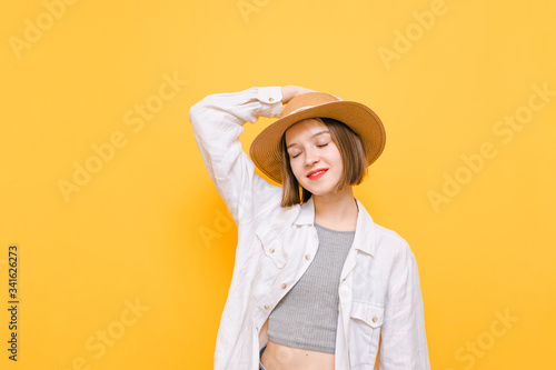 Portrait of smiling lady in hat and light clothes stands against yellow background with closed eyes and enjoys happy face, touches hat. Isolated. Copy space