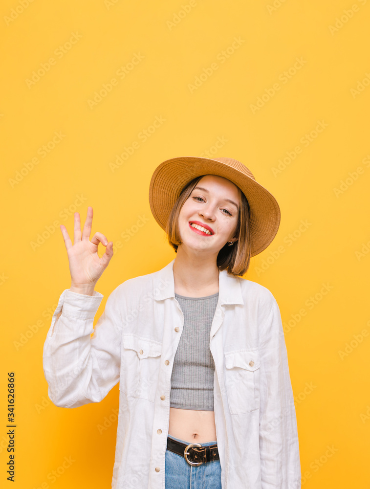 Attractive lady in hat and light clothing against yellow background, looking into camera with smile on face and showing gesture of approx. Happy female tourist shows ok gesture and laughs. Vertical Attractive lady in hat and light clothing against yellow background, looking into camera with smile on face and showing gesture of approx. Happy female tourist shows ok gesture and laughs. Vertical