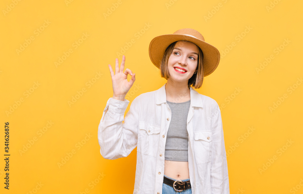 Attractive lady with happy face isolated on yellow background, looks away and shows OK gesture, wears summer clothes and hats. Girl tourist in hat on vacation, stands on yellow background. Attractive lady with happy face isolated on yellow background, looks away and shows OK gesture, wears summer clothes and hats. Girl tourist in hat on vacation, stands on yellow background.