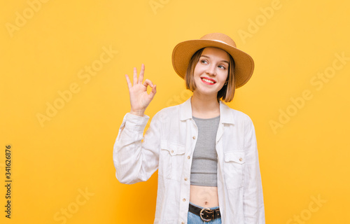 Attractive lady with happy face isolated on yellow background, looks away and shows OK gesture, wears summer clothes and hats. Girl tourist in hat on vacation, stands on yellow background.