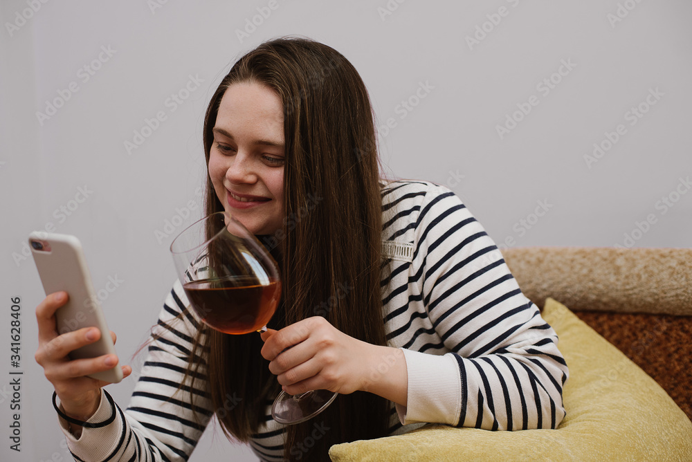 Close up photo of casual surprised young woman siting on the sofa and chatting maging video call on pone with friends while drinking wine