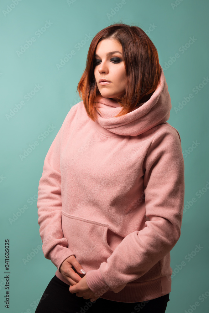 A girl in a pink hoodie looks down on a blue background. Studio woman portrait. A girl in a pink hoodie looks down on a blue background. Studio woman portrait.