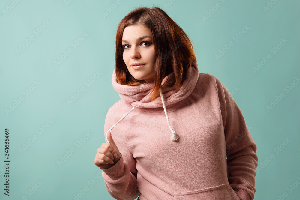 Redhead girl in a pink sweatshirt with a hood in an urban style smiles on a blue background. Redhead girl in a pink sweatshirt with a hood in an urban style smiles on a blue background.