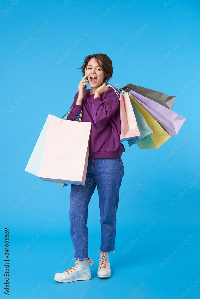 Good looking young joyful curly brunette woman having nice phone conversation and rejoicing about something, holding shopping bags while standing over blue background Good looking young joyful curly brunette woman having nice phone conversation and rejoicing about something, holding shopping bags while standing over blue background
