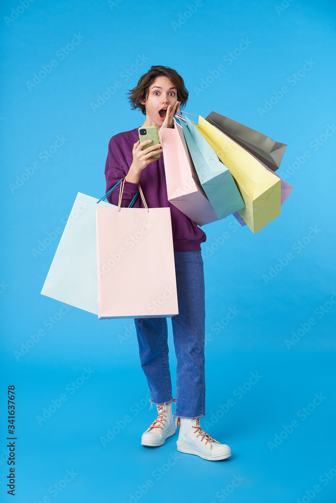 Amazed young brown-eyed curly brunette lady holding palm on her cheek while looking surprisedly at camera, keeping smartphone and purchases in raised hands while isolated over blue background Amazed young brown-eyed curly brunette lady holding palm on her cheek while looking surprisedly at camera, keeping smartphone and purchases in raised hands while isolated over blue background