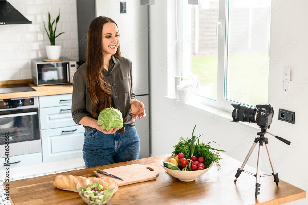 Young woman is recording video cooking lessons. She is preparing a salad in a cozy kitchen and taking pictures of herself on camera.
