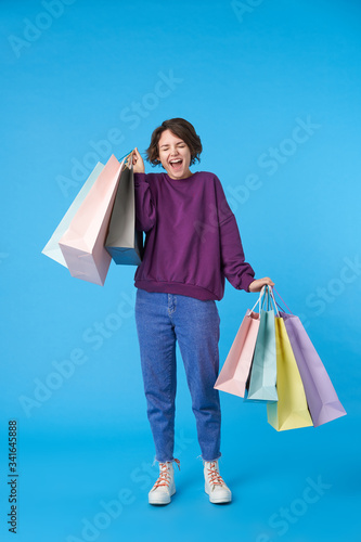 Good looking young curly brunette woman rejoicing with closed eyes while making shopping, holding paper bags while standing over blue background in casual clothes