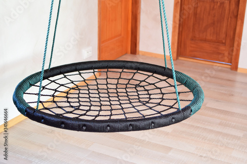 Rope circle green swing seat on light wall, wooden doors and laminate floor background. Black rope web nest for swinging closeup in empty room. Indoors leisure activities concept. 