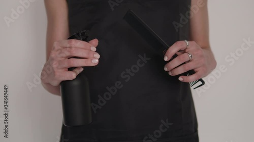Detail shot of hairdresser woman's hands holding and demonstrating professional barber tools comb and spray dispenser with black apron background on hairstyling salon 