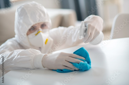 Woman in white workwear and protective gloves sparying the antiseptic liquid on the table