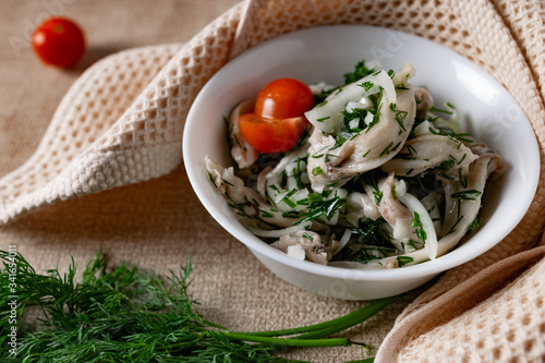 Marinated oyster mushrooms, garnished with cherry tomatoes and green dill. A dish with onions and butter in a white plate with a beige light cloth on the table