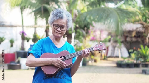 Portrait of an elderly woman playing ukulele in her garden. Relaxing by singing and play small guitar happy and enjoy life after retired. Concept of old people and health care