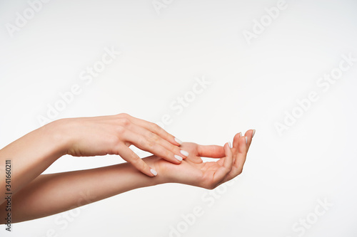 Beautiful young raised woman's hand with white manicure touching another one while moisturizing it with cream, isolated against white background