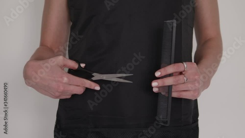 Detail shot of hairdresser woman's hands holding and demonstrating professional barber tools comb and scissors with black apron background on hairstyling salon 