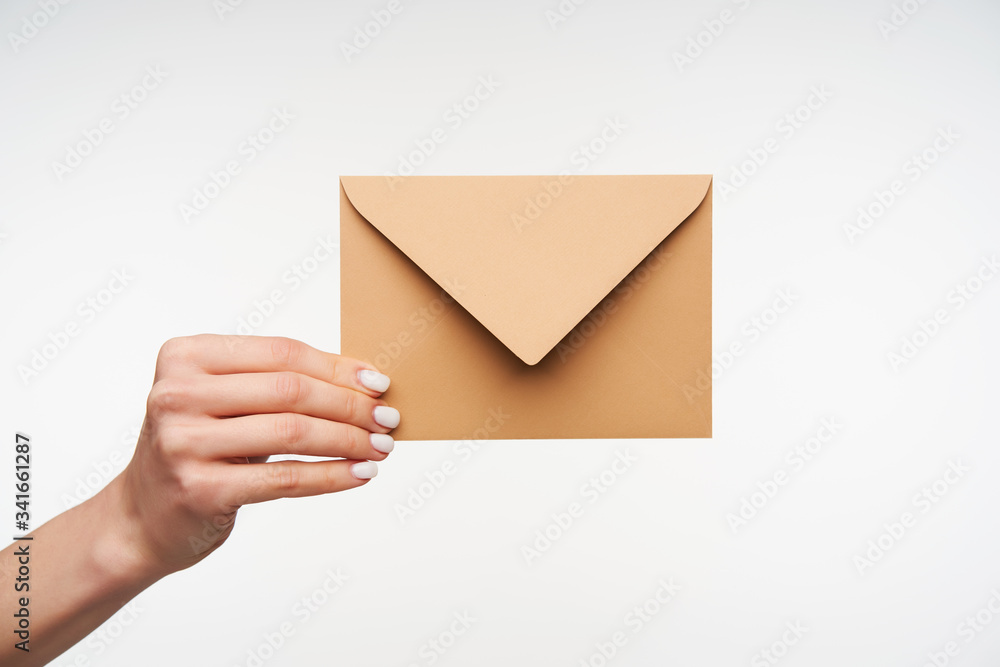 Cropped shot of raised young female's hand with white manicure holding craft envelope while being isolated against white background. Hands and gesturing concept