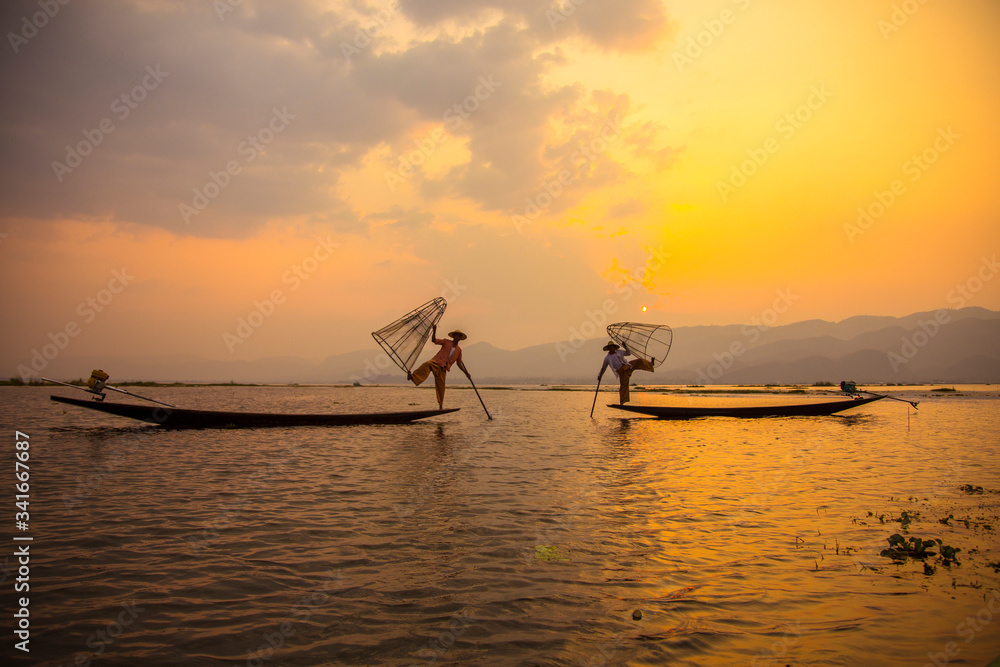 Inle Lake, Myanmar »; March 2018: Silhouette of two acrobatic fishermen in their boats with nets in the orange sunset on Inle Lake