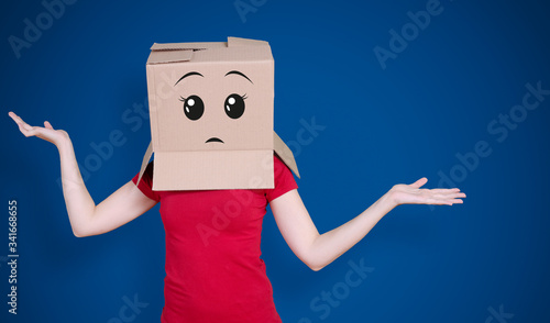 Person with cardboard box on its head and an expectant face expression stretching its arms out inquiringly on deep blue background photo