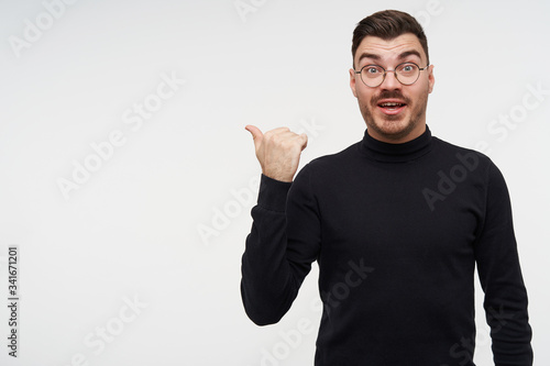 Agitated young lovely dark haired bearded man raising surprisedly eyebrows while showing aside with thumb, posing over white background in elegant wear © timtimphoto