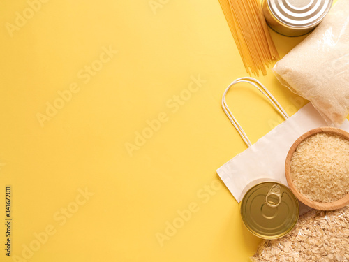 Dry goods, delivery, stockpiling, food supplies for staying home concept, donation, volunteer. Craft paper shopping bag, preserves, pasta, oatmeal, rice, sugar, on yellow background, copyspace