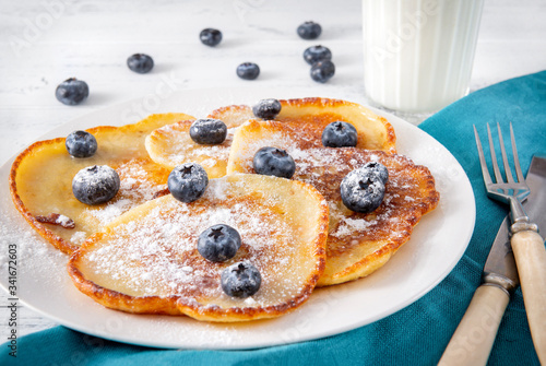 pancakes with  blueberries on a plate,  glass of milk,  fork,  knife on  blue napkin on a white wooden background