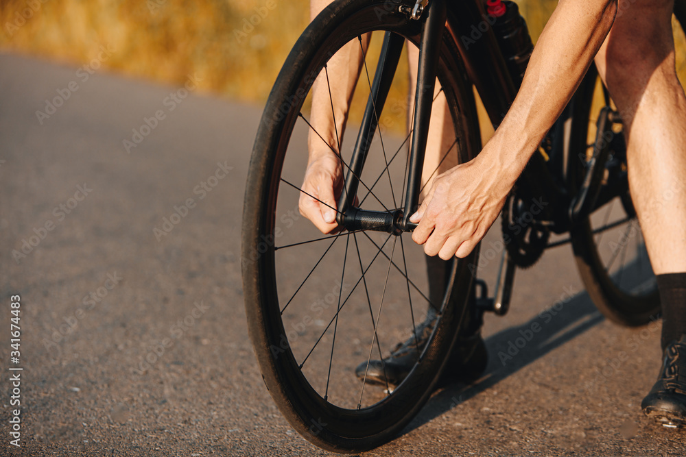 Close up of professional cyclist in activewear tighten nuts on bike wheel outdoors. Strong man repairing bike on paved road. Close up of professional cyclist in activewear tighten nuts on bike wheel outdoors. Strong man repairing bike on paved road.