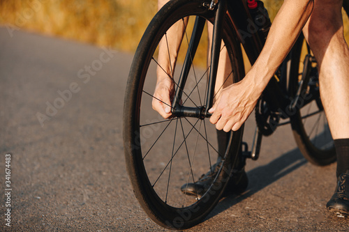 Close up of professional cyclist in activewear tighten nuts on bike wheel outdoors. Strong man repairing bike on paved road. © Tymoshchuk