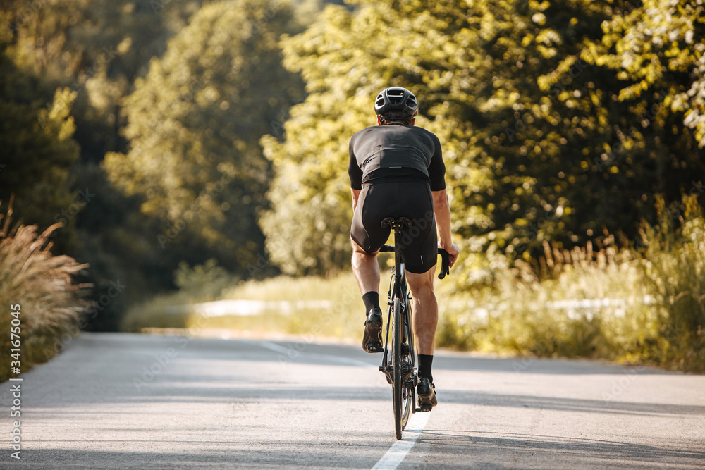 Obraz premium Back view of strong man in sport clothing and black helmet riding professional bike on nature. Mature sportsman enjoying favorite hobby on paved road.