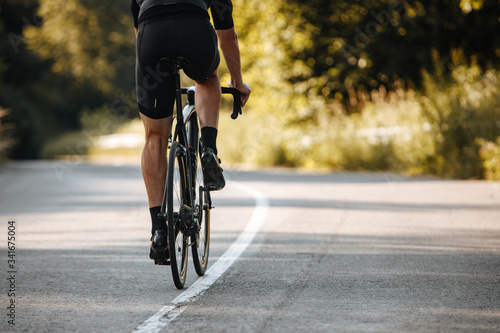 Back view of cyclist in activewear riding professional bike on paved road with blur background of green plants. Concept of active lifestyle and hard training