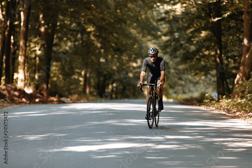 Full length portrait of bearded sportsman in black clothes, helmet and eyeglasses doing cycling workout on paved road among beautiful nature. Concept of active lifestyle.