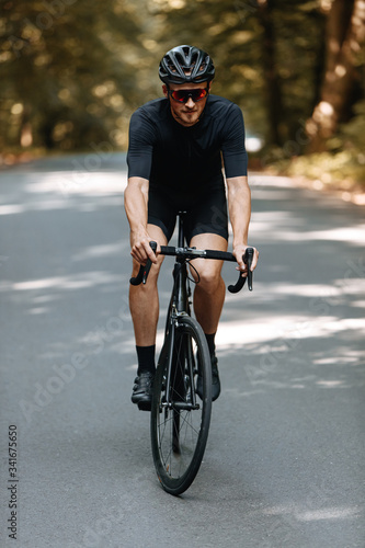Fototapeta Naklejka Na Ścianę i Meble -  Front view of strong male cyclist in sportswear riding black bike with blur background. Bearded man in protective helmet and eyeglasses improving skills and getting ready for competition
