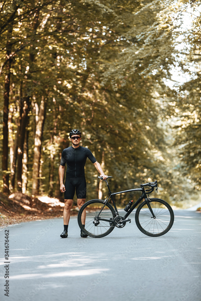 Confident bearded cyclist in special sport outfit standing near his black bike and looking at camera. Mature man in protective helmet and mirrored glasses enjoying workout outdoors.