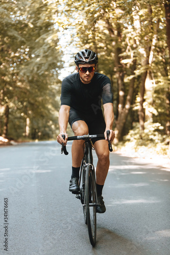 Front view of bearded man with athletic body shape dressed in sport clothing and black helmet biking in forest. Sporty person in mirrored eyeglasses enjoying summer nature during workout.