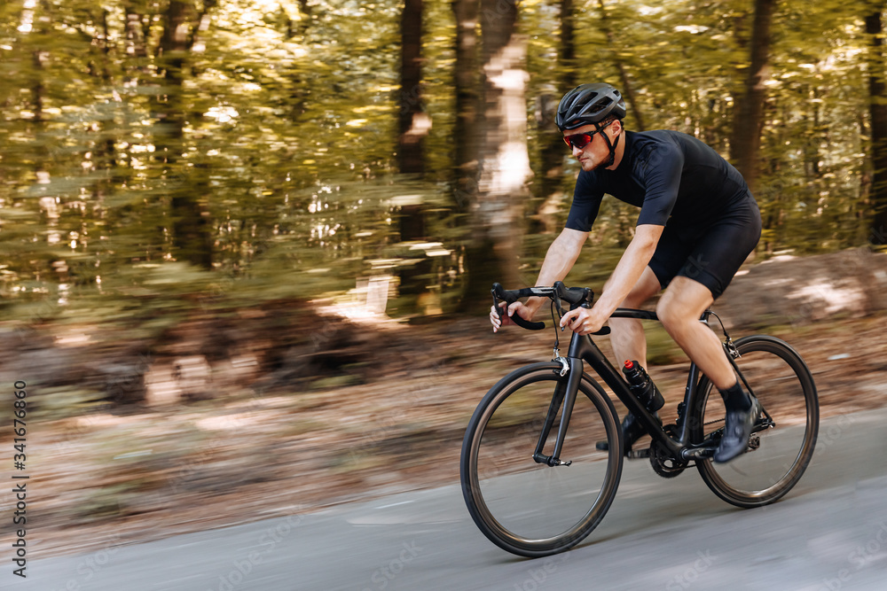 Fototapeta premium Motion blur of bearded cyclist wearing sport clothing, black helmet and mirrored glasses training with bike on asphalt road. Strong man improving skills and getting ready for competition