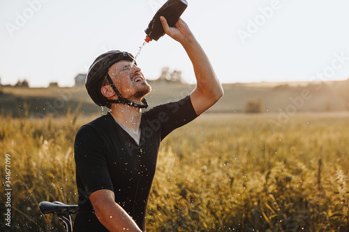 Mature tired athlete in sportswear and protective helmet splashing cold water on his face from black bottle after riding bike during sunny days. Bearded man refreshing after workout outdoors. photo