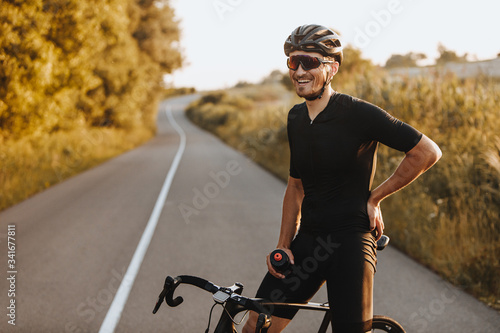 Cheerful bearded man in activewear, black helmet and sport glasses sitting on bike and looking on camera with beautiful nature around. Concept of active and healthy lifestyle.