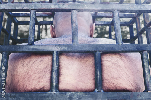 The bald man in the cage pressed his back against the iron bars. concept of prison and imprisonment