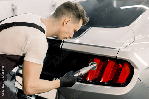 Detailing and polishing of car tail light on car. Young professional concentrated Caucasian male worker with orbital polisher in auto service polishing car taillights of white luxury car