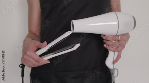 Detail shot of hairdresser woman's hands holding and demonstrating professional barber tools curling iron and hair dryer with black apron background on hairstyling salon 
