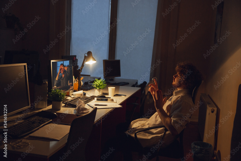 Online conference. Man working in office alone during COVID-19 quarantine, staying to late night. Young businessman, manager doing tasks with smartphone, laptop, tablet in empty workspace. Online conference. Man working in office alone during COVID-19 quarantine, staying to late night. Young businessman, manager doing tasks with smartphone, laptop, tablet in empty workspace.